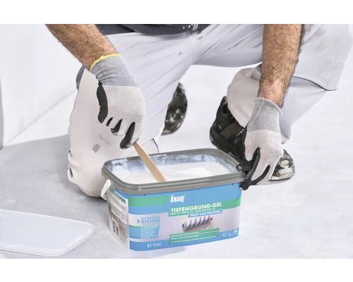 Une personne s'agenouille et mélange le gel de fond blanc Knauf dans un seau bleu avec un bâton en bois.