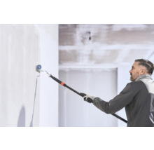 Un homme peint un mur avec un rouleau à peinture sur une perche télescopique dans une pièce lumineuse.