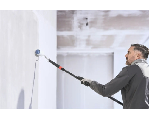 Un homme peint un mur avec un rouleau à peinture sur une perche télescopique dans une pièce lumineuse.