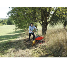 Un homme utilise une motofaucheuse dans une prairie avec de l'herbe haute sous un arbre.