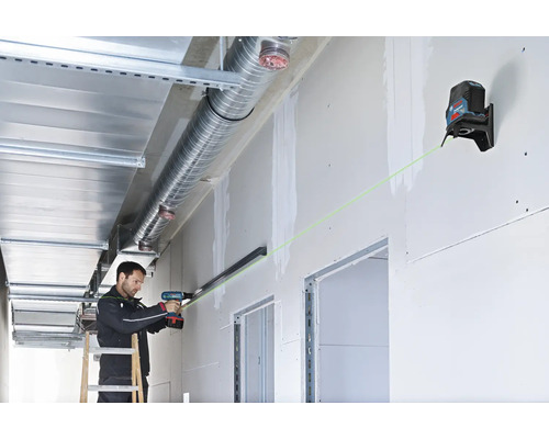 Un artisan utilise un laser en croix Bosch avec faisceau vert pour aligner un rail sur une cloison sèche avec une perceuse.