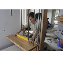 Artisan alignant une marche d'escalier en bois avec un niveau à bulle près d'un garde-corps métallique dans une pièce lumineuse en chantier.