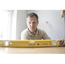 Un artisan regarde un niveau à bulle jaune Stabila type R 300 avec une longueur de 61 centimètres sur une table dans une pièce lumineuse.