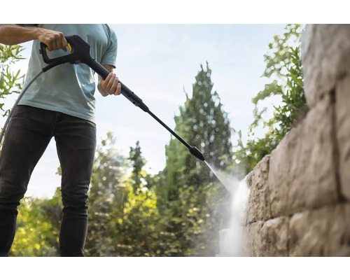 Eine Person reinigt eine strukturierte Steinmauer mit der Sprühlanze eines Hochdruckreinigers im Garten.