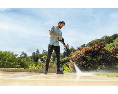 Ein Mann reinigt eine helle Steinterrasse im Garten mit einem Hochdruckreiniger bei sonnigem Wetter.