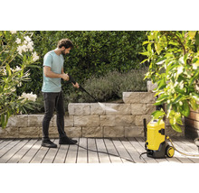 Ein Mann reinigt eine Steinmauer im Garten mit einem gelben Hochdruckreiniger auf einer Holzterrasse umgeben von Pflanzen.