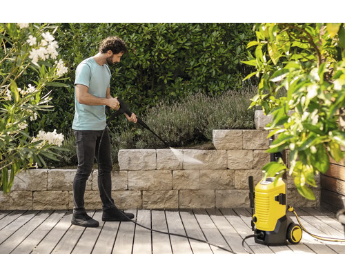 Ein Mann reinigt eine Steinmauer im Garten mit einem gelben Hochdruckreiniger auf einer Holzterrasse umgeben von Pflanzen.