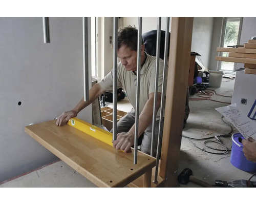 Artisan alignant une marche d'escalier en bois avec un niveau à bulle près d'un garde-corps métallique dans une pièce lumineuse en chantier.