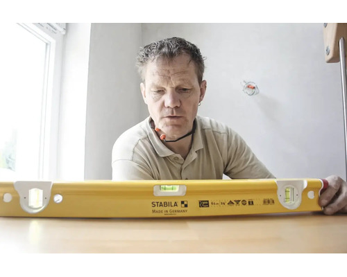 Un artisan regarde un niveau à bulle jaune Stabila type R 300 avec une longueur de 61 centimètres sur une table dans une pièce lumineuse.
