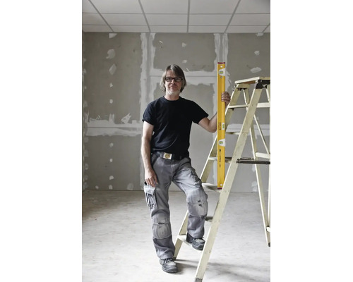 Artisan en pantalon de travail gris et t-shirt noir debout près d'une échelle avec un niveau à bulle jaune dans une pièce en chantier.