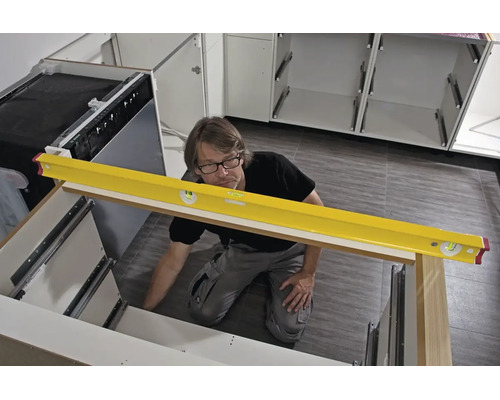 Un homme utilise un niveau à bulle jaune pour aligner des caissons de cuisine blancs lors de l'installation sur un sol carrelé.