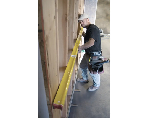 Un ouvrier du bâtiment utilise un long niveau à bulle jaune pour vérifier l'alignement d'une cloison en bois sur un chantier.