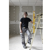 Artisan en pantalon de travail gris et t-shirt noir debout près d'une échelle avec un niveau à bulle jaune dans une pièce en chantier.
