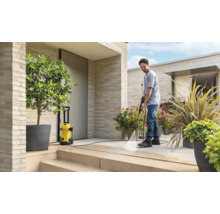 Un homme nettoie une terrasse moderne en pierre claire avec un nettoyeur haute pression jaune. L'espace extérieur est lumineux avec des plantes.