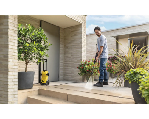 Un homme nettoie une terrasse moderne en pierre claire avec un nettoyeur haute pression jaune. L'espace extérieur est lumineux avec des plantes.