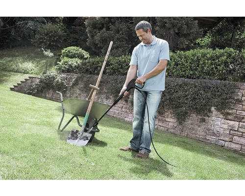 Un homme nettoie une pelle sale dans une brouette a l aide d un nettoyeur haute pression sur une pelouse dans un jardin.