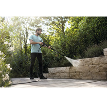 Un homme nettoie un mur en pierre naturelle sur une terrasse en bois avec un nettoyeur haute pression dans un jardin lumineux et ordonné.
