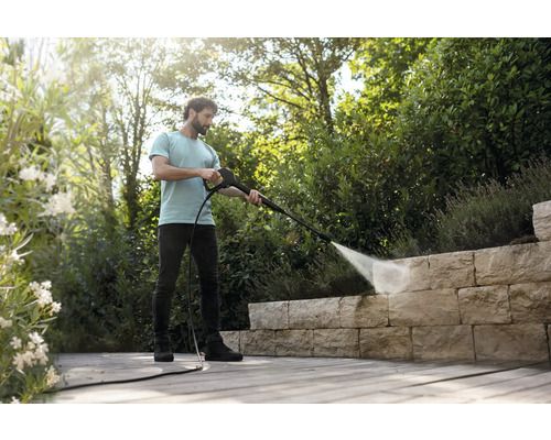 Un homme nettoie un mur en pierre naturelle sur une terrasse en bois avec un nettoyeur haute pression dans un jardin lumineux et ordonné.