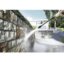 Nettoyage d'un mur en pierre naturelle dans un jardin lumineux avec un nettoyeur haute pression, à côté d'escaliers et de plantes.