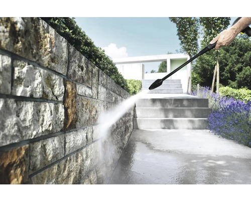 Nettoyage d'un mur en pierre naturelle dans un jardin lumineux avec un nettoyeur haute pression, à côté d'escaliers et de plantes.