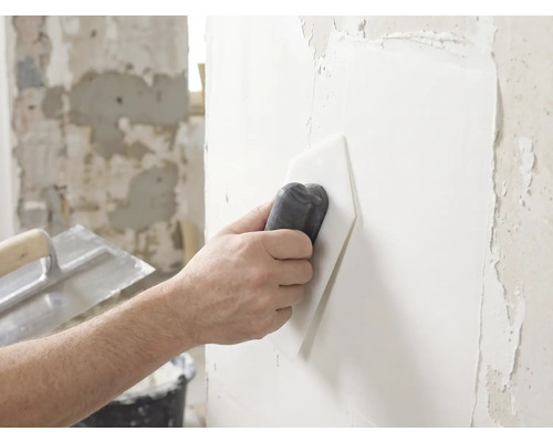 Une main lisse de l'enduit blanc sur un mur à l'aide d'un lisseur en plastique.