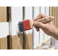 Une main peint une clôture en bois avec un pinceau plat Rautner et de la peinture blanche.