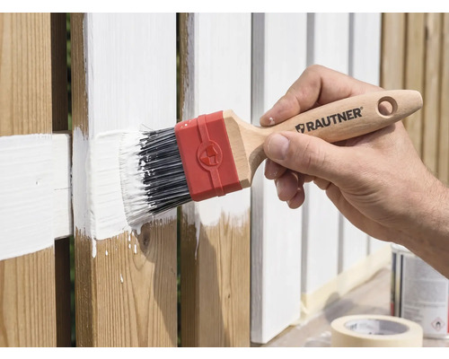 Une main peint une clôture en bois avec un pinceau plat Rautner et de la peinture blanche.