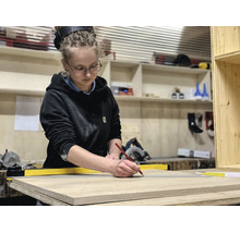 Une femme dans un atelier marque une planche de bois avec un crayon et une règle jaune. Des étagères avec des outils sont en arrière-plan.