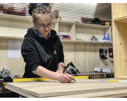 Une femme dans un atelier marque une planche de bois avec un crayon et une règle jaune. Des étagères avec des outils sont en arrière-plan.