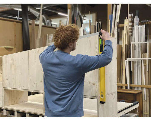 Personne vérifiant l'alignement vertical d'un meuble en bois avec un niveau à bulle numérique dans un atelier.