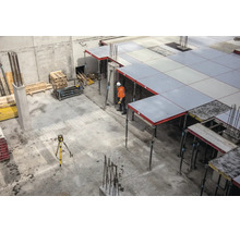 Chantier avec coffrage de dalle soutenu par des étais, un ouvrier en tenue orange et un niveau laser sur un trépied.