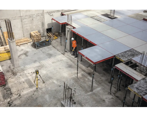 Chantier avec coffrage de dalle soutenu par des étais, un ouvrier en tenue orange et un niveau laser sur un trépied.