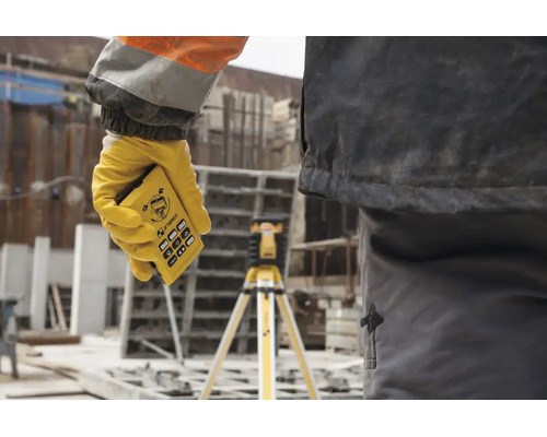 Main tenant une télécommande Stabila pour laser rotatif sur un chantier, avec un laser sur trépied en arrière-plan.
