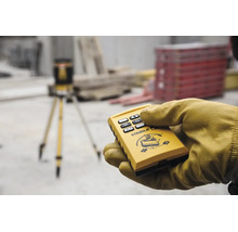 Main gantée tenant une télécommande pour laser rotatif Stabila sur un chantier avec trépied en arrière-plan.