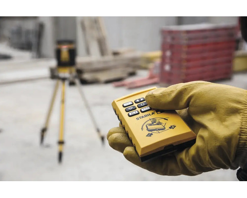 Main gantée tenant une télécommande pour laser rotatif Stabila sur un chantier avec trépied en arrière-plan.