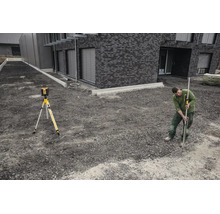 Un homme utilise un laser rotatif sur un trépied et une mire avec récepteur sur un chantier devant un bâtiment moderne en pierre.