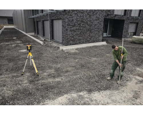 Un homme utilise un laser rotatif sur un trépied et une mire avec récepteur sur un chantier devant un bâtiment moderne en pierre.