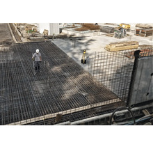 Ouvrier sur un chantier avec un ferraillage étendu en treillis soudés et un appareil de mesure sur un trépied.