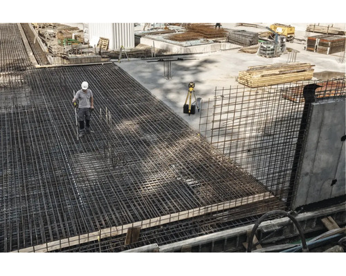 Ouvrier sur un chantier avec un ferraillage étendu en treillis soudés et un appareil de mesure sur un trépied.