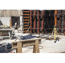Chantier avec laser rotatif Stabila sur trépied, murs de coffrage en métal, scie circulaire sur table et un ouvrier du bâtiment.