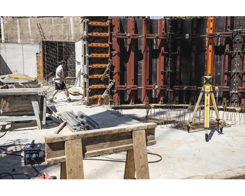 Chantier avec laser rotatif Stabila sur trépied, murs de coffrage en métal, scie circulaire sur table et un ouvrier du bâtiment.