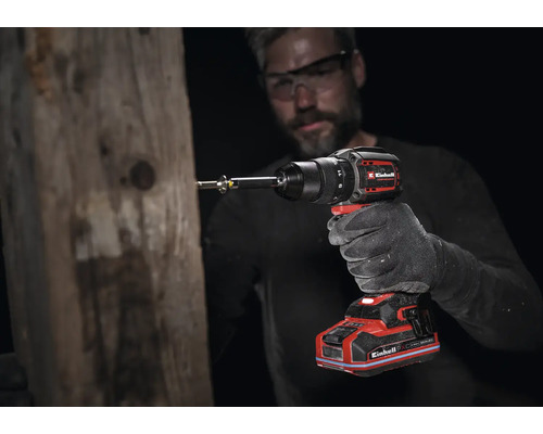 Perceuse-visseuse sans fil Einhell utilisée sur une poutre en bois par une personne portant un gant de travail et des lunettes de sécurité.