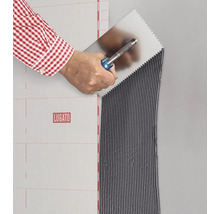 Eine Hand trägt mit einer Zahnspachtel Fliesenkleber auf eine Wand neben einer Lugato Abdichtungsbahn auf.