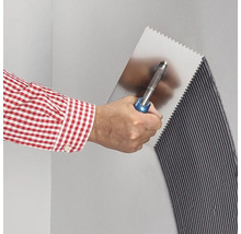 Eine Hand trägt mit einer Zahnkelle Fliesenkleber auf eine Wand auf, wodurch ein gleichmäßiges Rillenmuster entsteht.