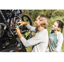 Un homme attache un VTT à un porte-vélos sur une voiture avec un cadenas et un câble antivol, tandis qu'une femme regarde.