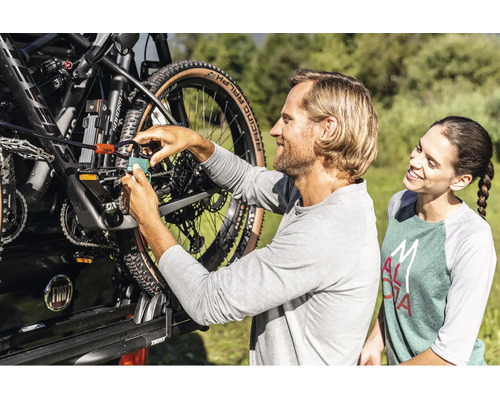 Un homme attache un VTT à un porte-vélos sur une voiture avec un cadenas et un câble antivol, tandis qu'une femme regarde.