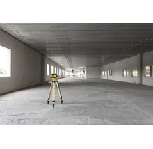 Laser rotatif sur un trépied dans un grand bâtiment industriel brut et lumineux avec sol en béton et hauts plafonds.