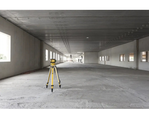 Laser rotatif sur un trépied dans un grand bâtiment industriel brut et lumineux avec sol en béton et hauts plafonds.