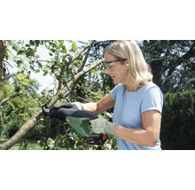 Femme coupant une branche dans un jardin avec une scie de jardin sans fil Bosch, portant des lunettes de protection et des gants.
