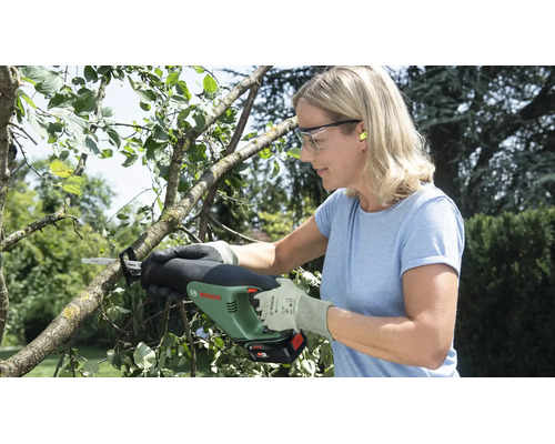 Femme coupant une branche dans un jardin avec une scie de jardin sans fil Bosch, portant des lunettes de protection et des gants.